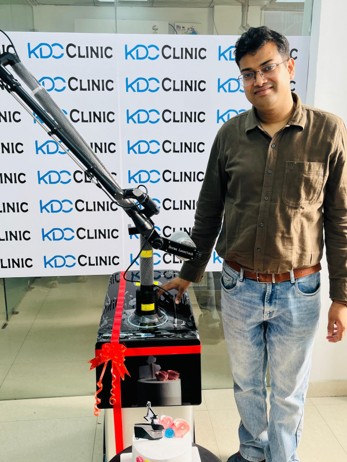 Man in a brown shirt and jeans stands beside a wheeled medical device with a red ribbon at a KDC Clinic backdrop.
