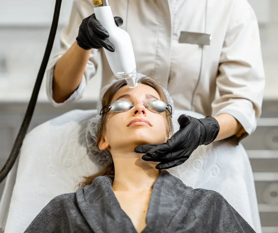 Medical professional performs a facial laser treatment on a woman wearing protective goggles and a robe; handheld device near her chin/cheek.