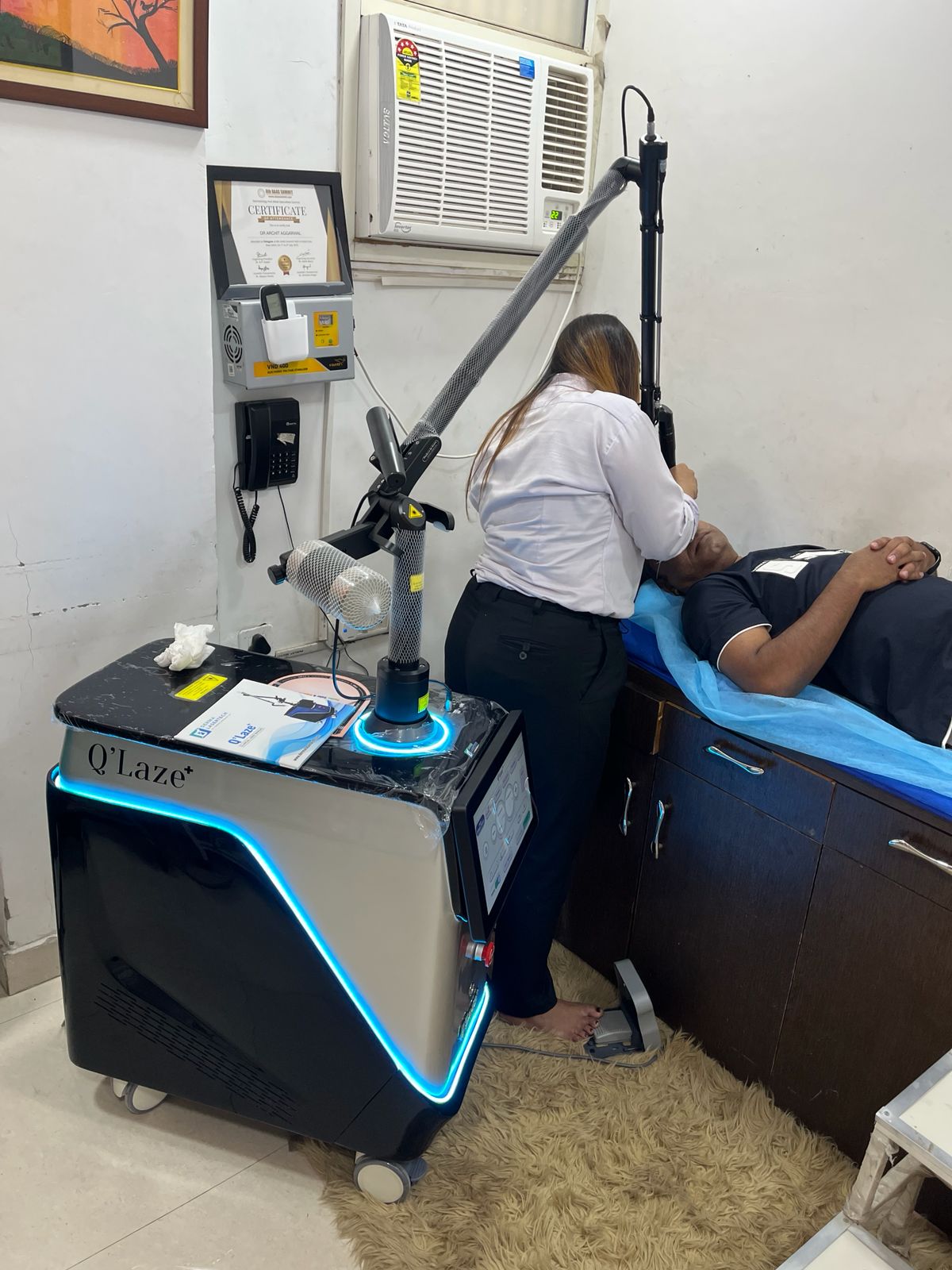 Patient lies on a treatment bed while a clinician uses a Q'Laze laser with a flexible arm for cosmetic treatment.