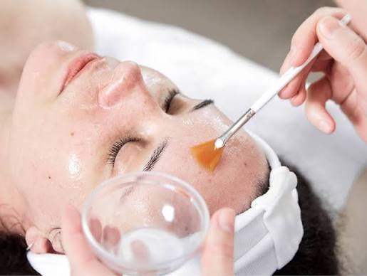 Esthetician applies an orange facial mask with a brush to a client’s cheek during a spa facial.